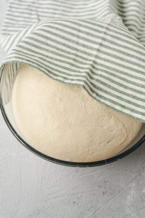 Fresh bread dough rising in a glass bowl with a striped tea towel on top.