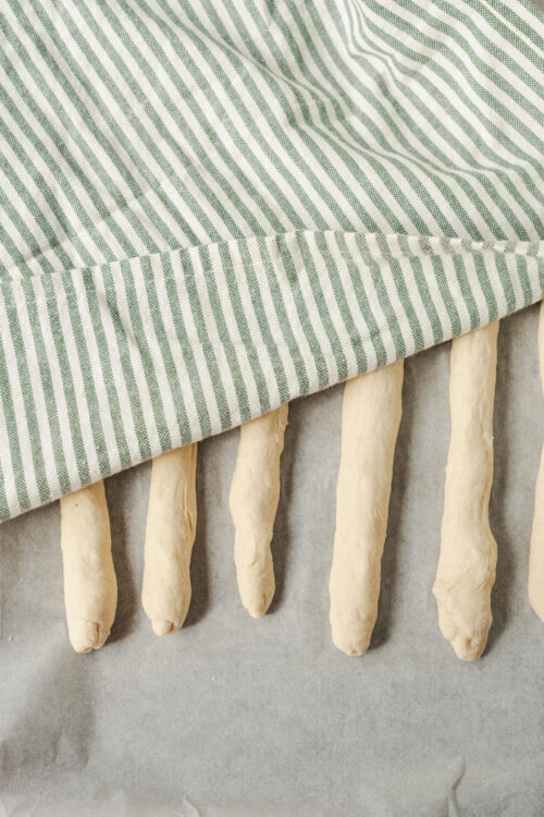 Rich butter cookie dough shaped into fingers, covered with green striped kitchen towel on baking sheet.