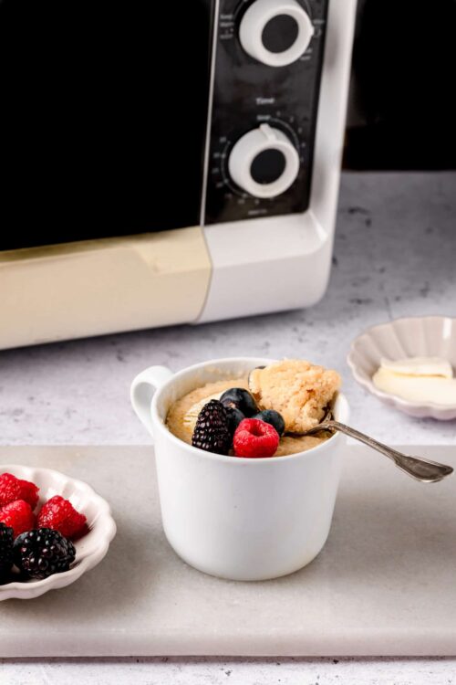 Creamy berry cobbler in a white mug with fresh raspberries, blackberries, and blueberries on a marble surface.