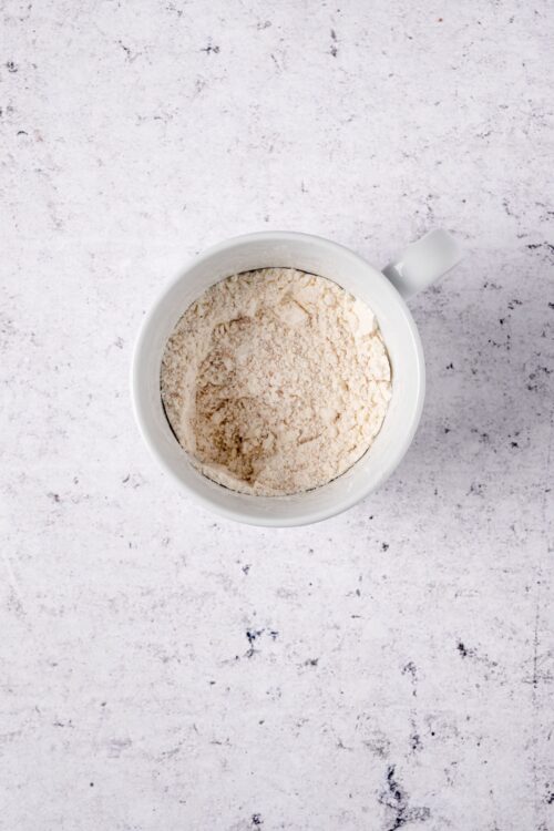 Flour in a white mug on a textured white surface for baking or cooking.