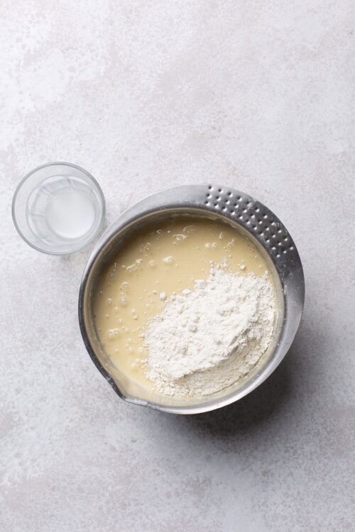 Flour and wet ingredients in mixing bowl for baking, on light textured surface.