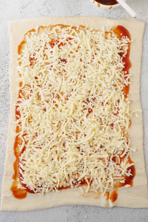 Cheese pizza preparation with shredded cheese on dough, tomato sauce, and baking ingredients.