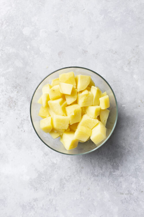 Diced raw potatoes in a clear glass bowl on a gray textured surface.