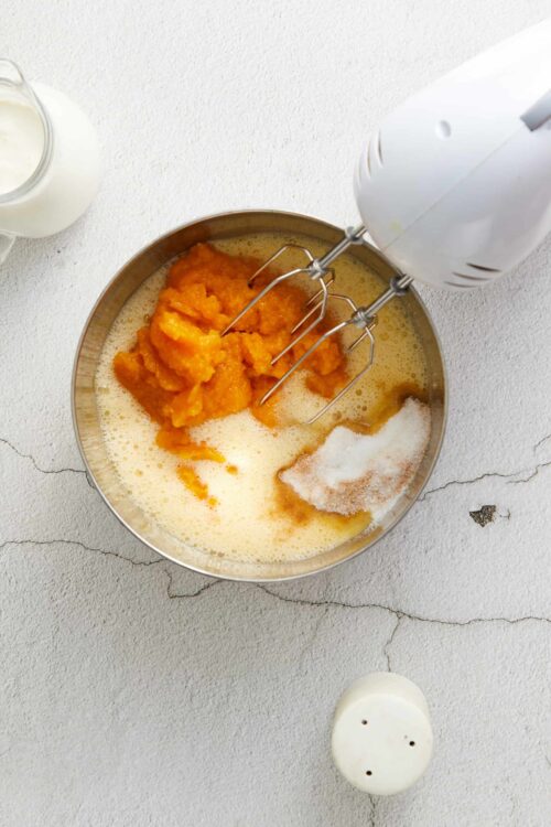 Sweet potato and sugar mixture in a mixing bowl with hand mixer attachments on a textured white surface.