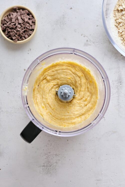 Creamy lemon cake batter in a food processor with chocolate chips and oats nearby.