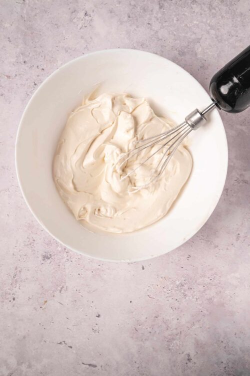 Cream cheese mixed in a bowl with a hand mixer on light pink textured background.