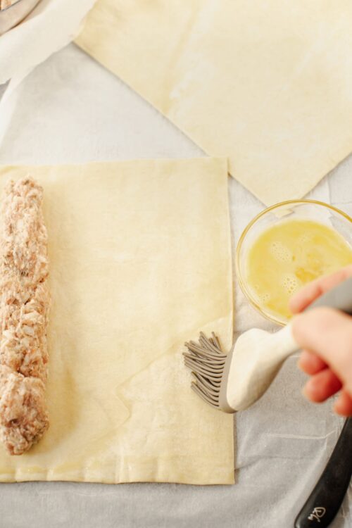 Butter and puff pastry ready for baking, with a hand using a pastry brush, on parchment paper.