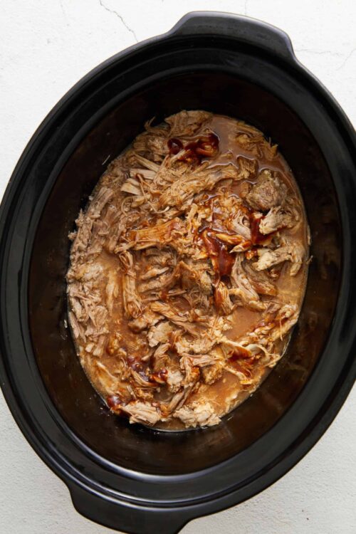 Shredded slow cooker pulled pork with barbecue sauce in a black crockpot.