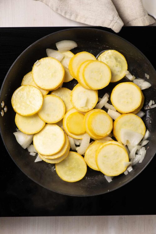 Thinly sliced yellow squash and onions cooking in a skillet.