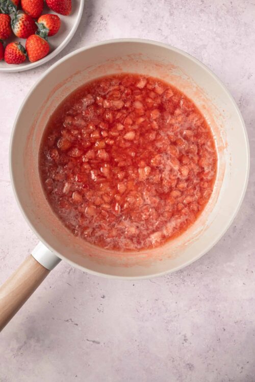 Fresh strawberry jam cooking in a white saucepan on a pink surface.