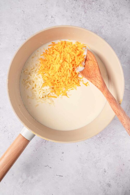 Creamy cheese sauce with shredded cheddar and mozzarella in a beige saucepan on a light textured surface.