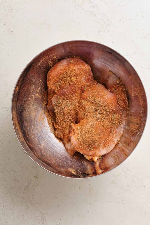 Seasoned raw chicken breasts in a metal bowl ready for baking or grilling.