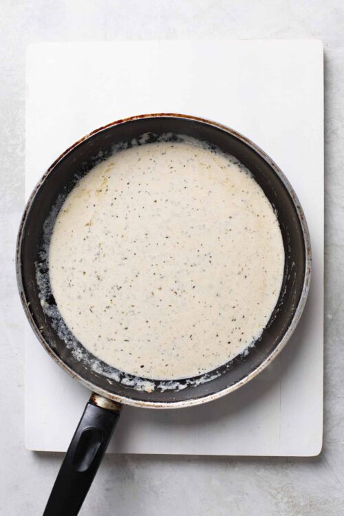 Creamy white sauce with black pepper flecks in a black skillet on a white cutting board.