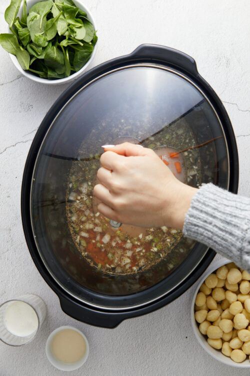 Steamy slow cooker with vegetables, herbs, and broth on a textured white surface.