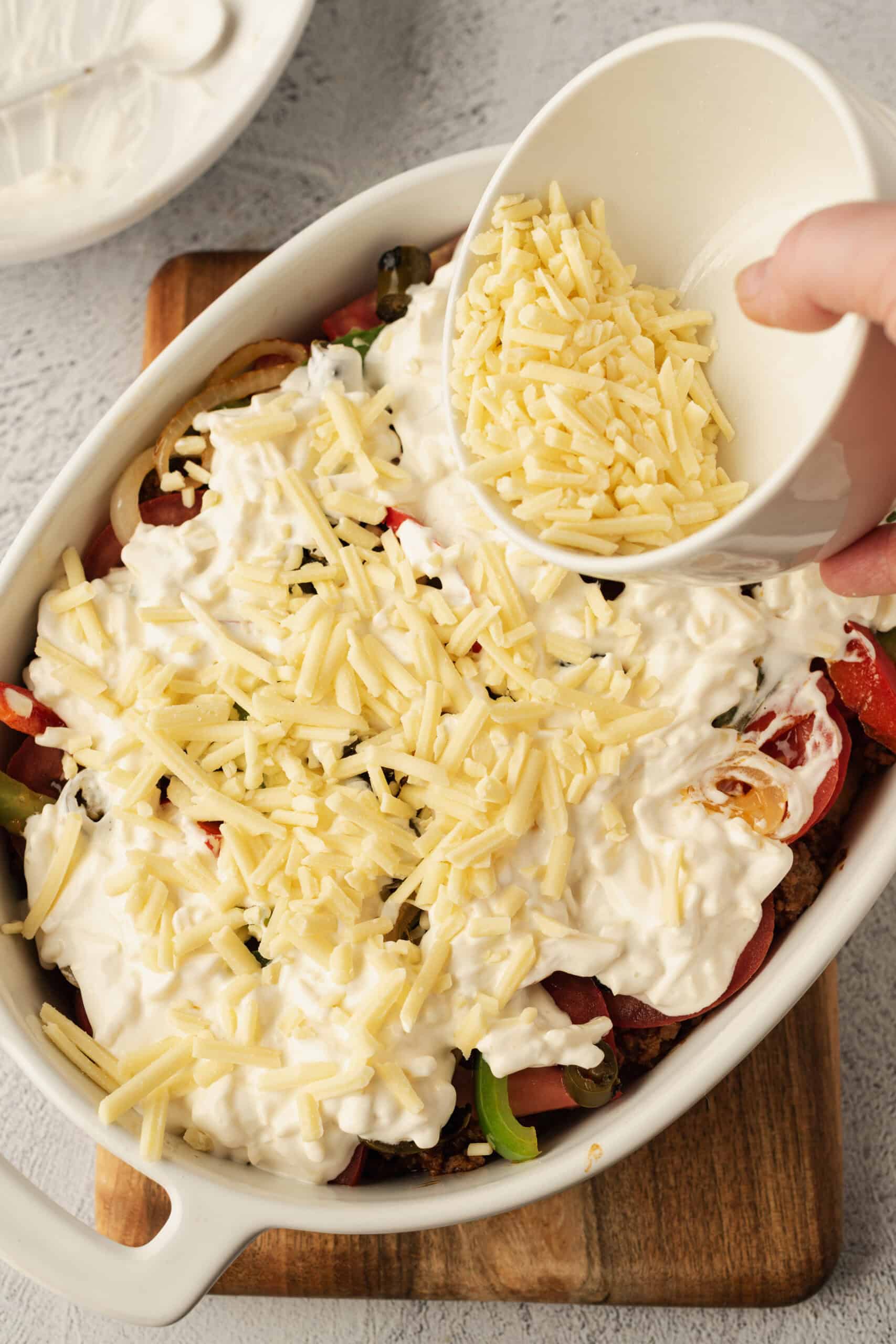 Shredded cheese being sprinkled onto a layered casserole dish with vegetables and sour cream.