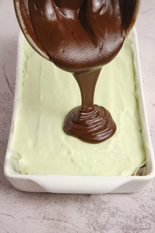 Decadent chocolate frosting being poured onto green custard dessert in a white dish.