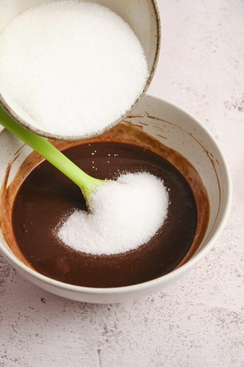 Fine granulated sugar being added to melted chocolate in a baking bowl.
