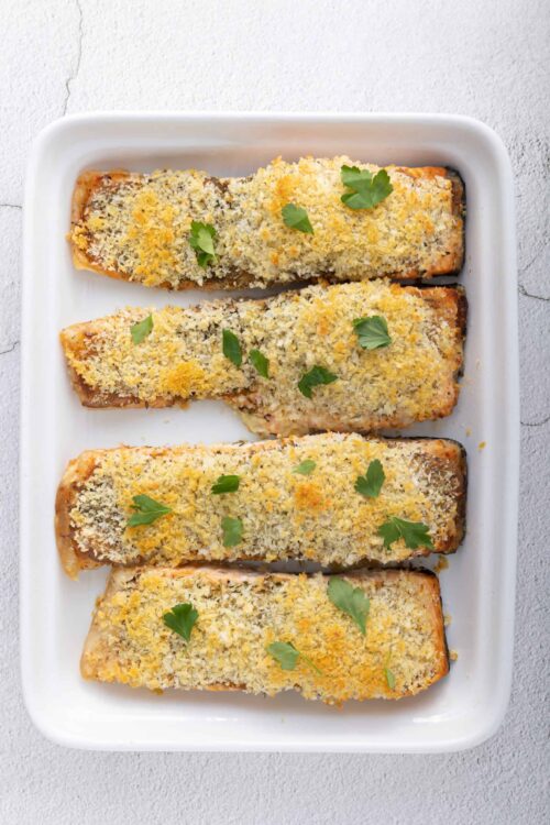 Baked salmon with breadcrumb topping and fresh herbs on white baking dish.