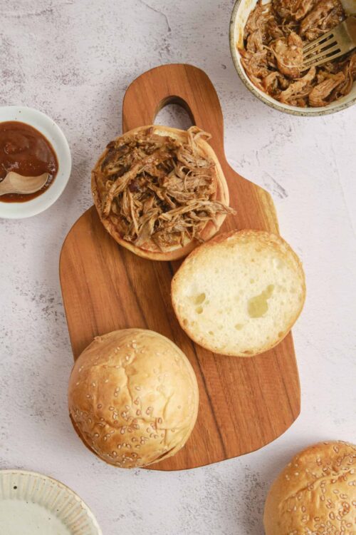 Shredded BBQ pulled pork sandwiches with soft hamburger buns on a wooden serving board.