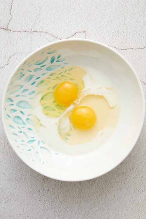 Creamy cracked eggs in a decorative white bowl with cutout floral design, on a textured grey surface.