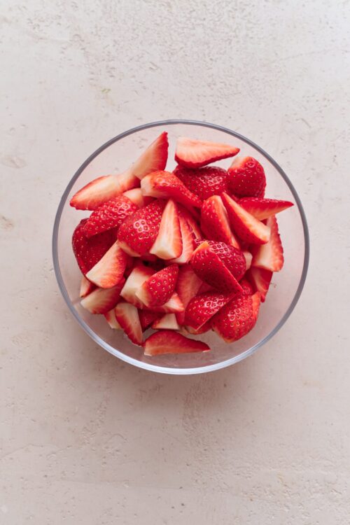 Fresh sliced strawberries in a clear glass bowl on a neutral background.