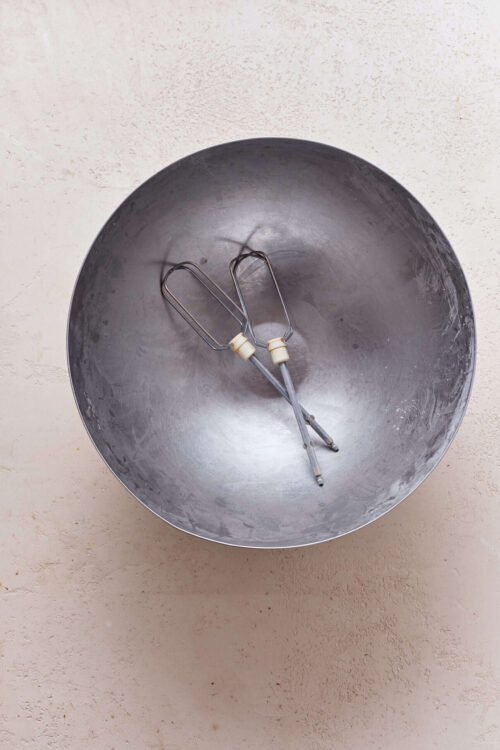 Mixing bowl with hand mixer beaters for baking desserts or bread, on a textured surface.
