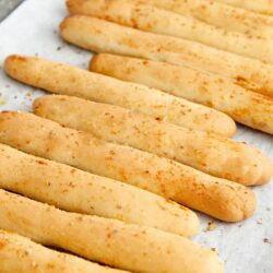 Golden baked breadsticks sprinkled with herbs and cheese on baking sheet.