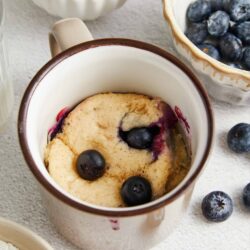Blueberry cobbler in a mug with fresh blueberries and baking ingredients around it.