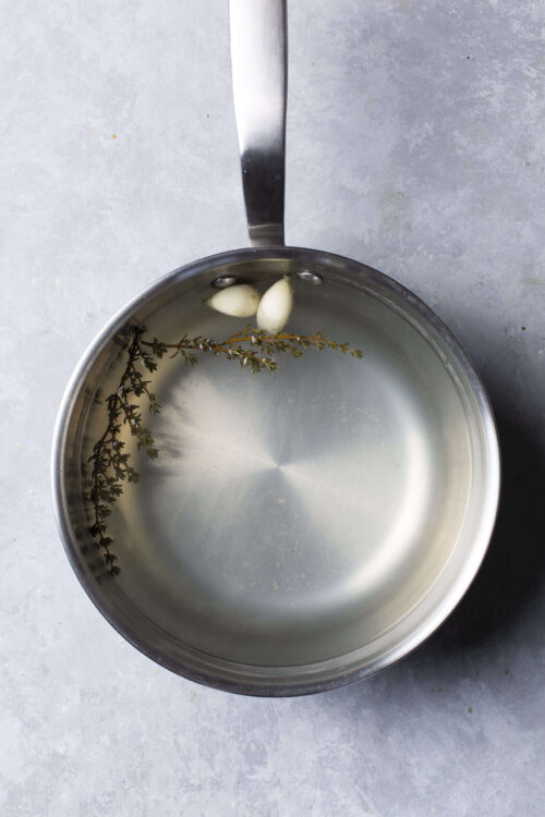 Bright stainless steel saucepan with garlic cloves and fresh thyme in water, minimalism, kitchen cooking essentials.