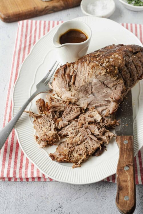 Succulent shredded beef roast with rich gravy, served on a white platter with carving knife and fork.