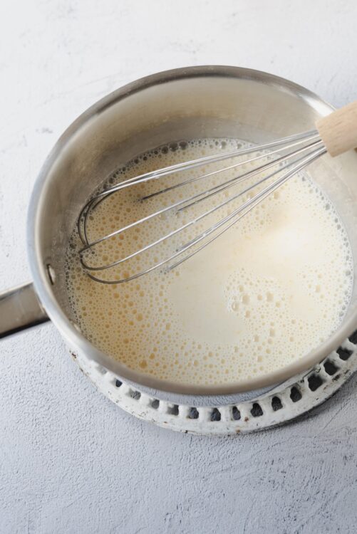 Cream mixture being whisked in a stainless steel saucepan on a stove.