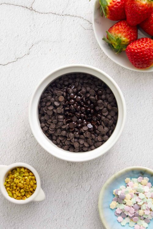 Fresh strawberries on white plate with chocolate chips and colorful sprinkles, ideal for baking or dessert decorating.