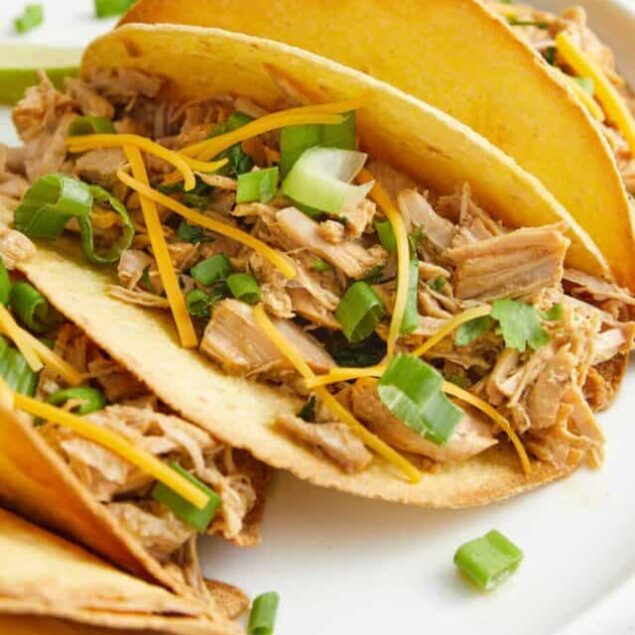 Shredded chicken tacos with cheese, green onions, and lemon wedges on a white plate, fresh Mexican-inspired recipe.