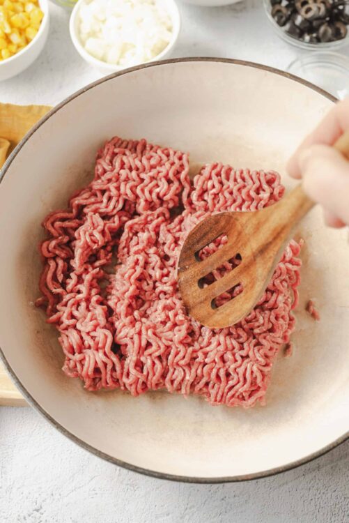 Raw ground beef in a skillet being cooked with a wooden spatula.