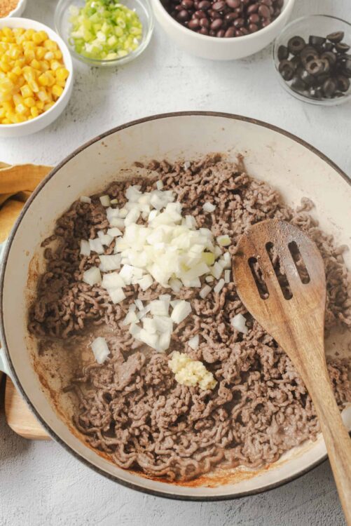 Ground beef cooking in a skillet with chopped onions and garlic, surrounded by bowls of corn, olives, celery, and black beans.