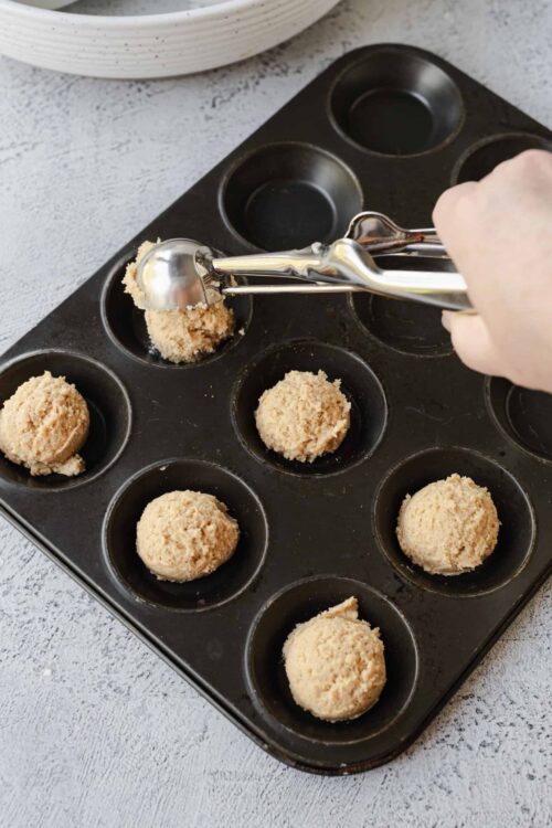 Soft cookie dough balls being scooped into a non-stick muffin tin for baking.
