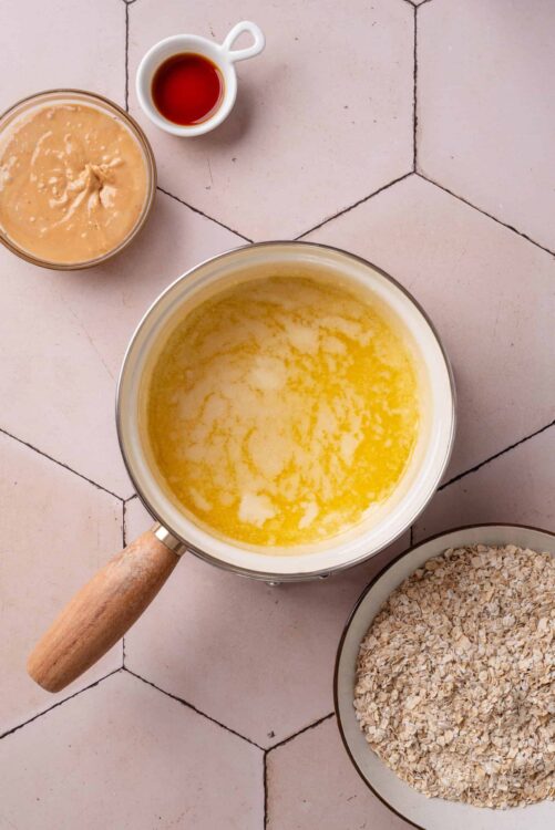 Creamy oatmeal with butter in a saucepan on tiled surface, with oats, syrup, and brown sugar nearby.
