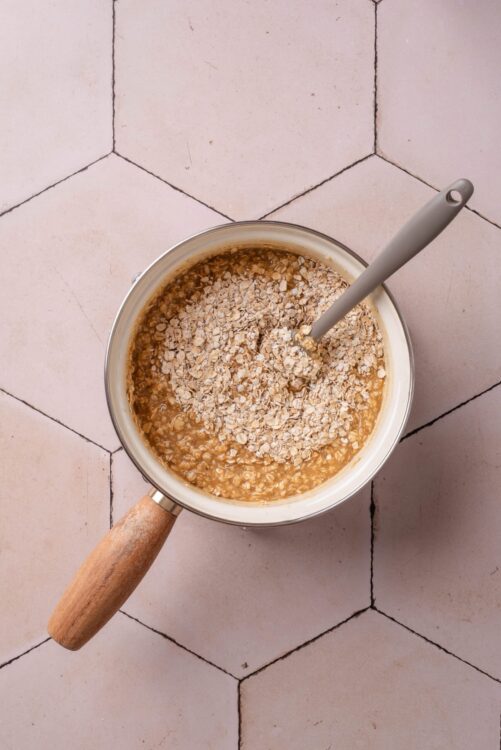 Creamy caramel mixture with oats in a saucepan on pink tiled surface.