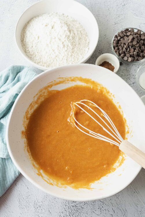 Creamy pumpkin cake batter with chocolate chips and baking ingredients on light surface.