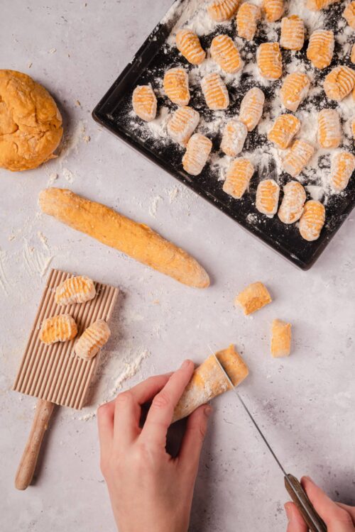 Fried gnocchi on a baking sheet dusted with flour, kitchen scene with a hand slicing cooked gnocchi.
