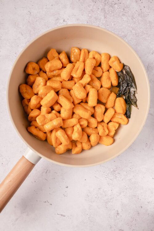 Creamy orange gnocchi with sage leaves in a beige skillet over a light textured surface.