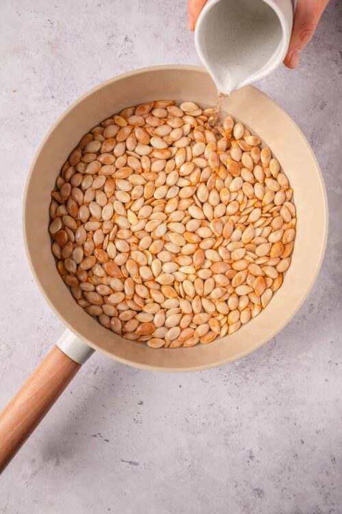 Roasted pumpkin seeds in a beige skillet with pouring of liquid.