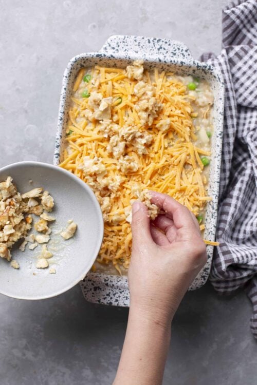 Cheesy chicken casserole being topped with crumbled cooked chicken pieces.