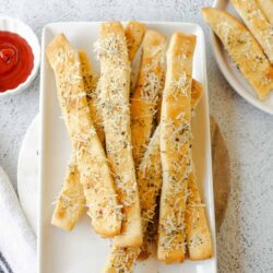 Golden cheesy breadsticks with grated cheese and herbs on a white platter.