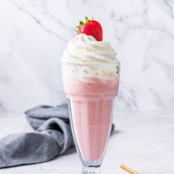 Fresh strawberry milkshake topped with whipped cream and a whole strawberry, served in a tall glass.