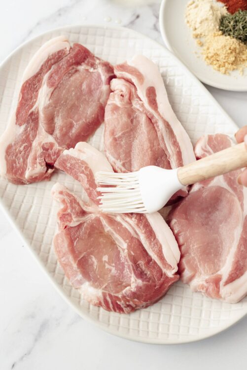 Raw pork chops on a white dish, being prepared for cooking.