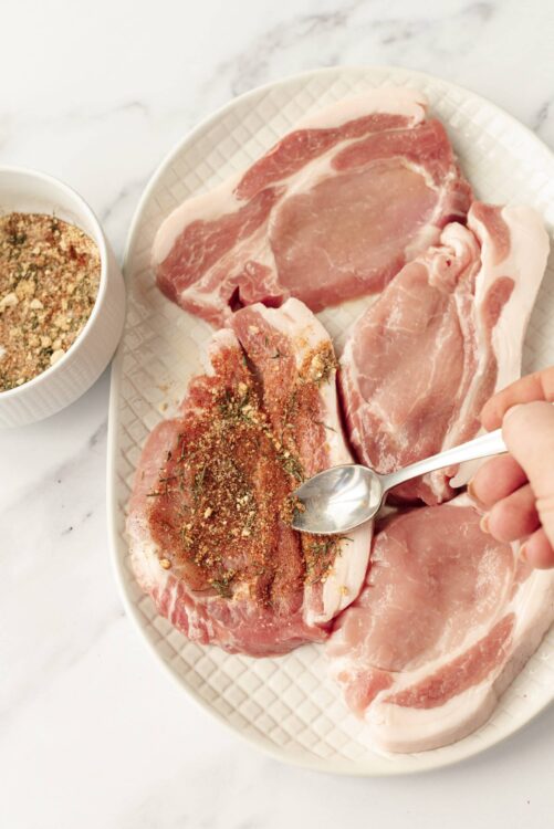 Seasoned raw pork chops for baking, with herbs and spices on a white plate.