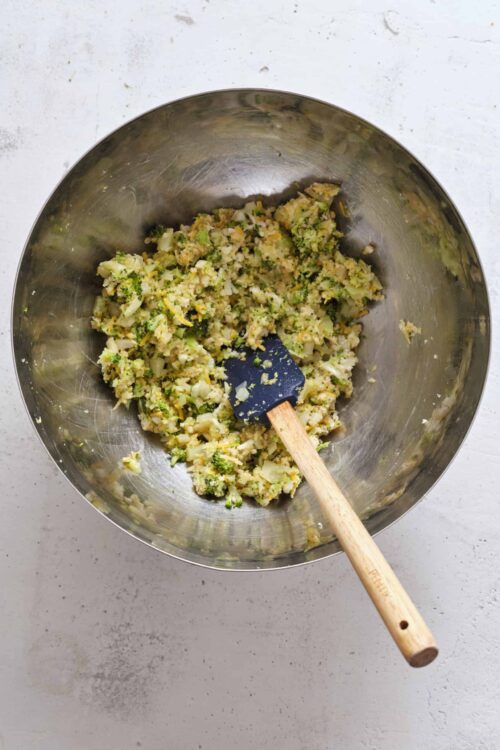 Mashed broccoli and cauliflower in stainless steel mixing bowl with spatula.