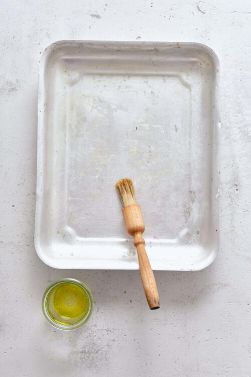 Bare baking sheet with pastry brush and jar of olive oil on white surface, plain baking tools for cooking or baking.
