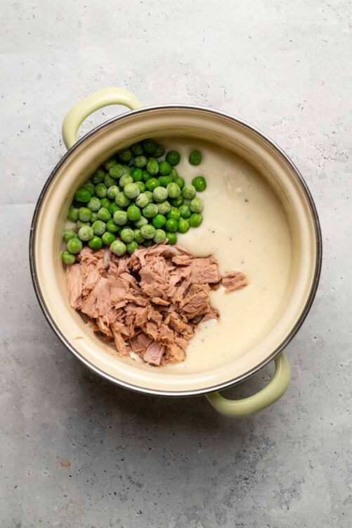 Fresh green peas, shredded tuna, and creamy sauce in a beige and green pot, ready for a delicious casserole.
